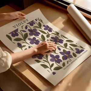 A person's hands are holding a poster with a floral design featuring purple flowers and green leaves.