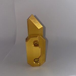 May include: A gold-colored metal object with a geometric design. The object has a sharp point at the top and a rectangular base.