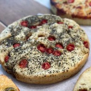 May include: Close-up of a round focaccia bread topped with red cherry tomatoes and herbs. The bread has a light, airy texture and is sprinkled with dark green herbs. Other pieces of bread are visible in the background.