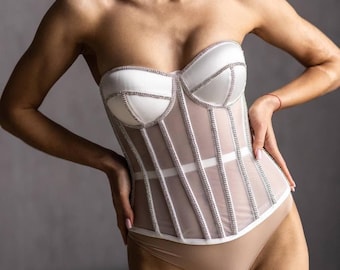 May include: A strapless white and nude corset with rhinestone detailing. The corset features a structured bodice with vertical boning and a sheer mesh panel. The garment is paired with a nude bodysuit.