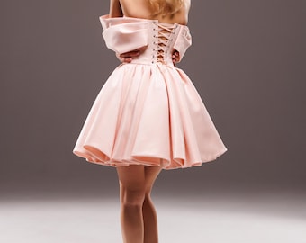 May include: A light pink, strapless mini dress with a corset back and a flared skirt. The dress is made of a smooth fabric and is paired with beige high heels. The model is facing away from the camera.