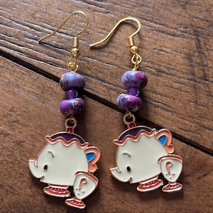 Peut inclure: Paire de boucles d'oreilles dorées avec un motif de théière. Les boucles d'oreilles ont des perles violettes et roses et un charm de théière blanc avec des accents rouges, bleus et oranges. Sur une surface en bois.