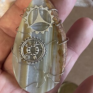Peut inclure: Un collier pendentif de style steampunk. Le pendentif est une pierre en forme de goutte d'eau avec des bandes crème, marron et beige. Deux breloques argentées en forme de roue dentée sont fixées à la pierre avec du fil d'argent. La chaîne du collier est également argentée.