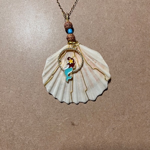 May include: A gold chain necklace with a seashell pendant. The seashell is white with a gold wire design and a small mermaid charm inside a gold circle.