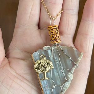 Peut inclure: Un collier en chaîne dorée avec un pendentif en agate grise enveloppé de fil d'argent. Le pendentif comprend également un charme d'arbre en or et une perle jaune et brune.