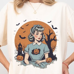 May include: A white t-shirt with a vintage illustration of a woman in a blue dress with a pumpkin in her chest. The illustration is set against a background of a full moon, bats, and a spooky tree.