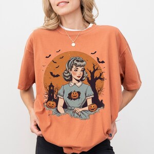 May include: A woman wearing a burnt orange t-shirt with a vintage illustration of a woman in a blue dress with pumpkins and bats. The t-shirt has a crew neck and short sleeves.