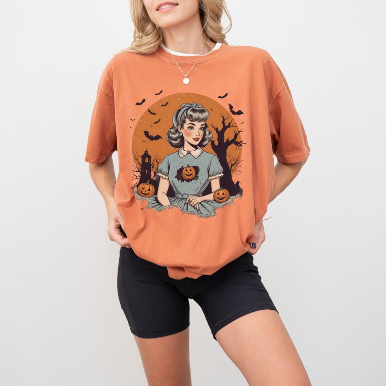 May include: A woman wearing a burnt orange t-shirt with a vintage illustration of a woman in a blue dress with pumpkins and bats. The t-shirt has a crew neck and short sleeves.