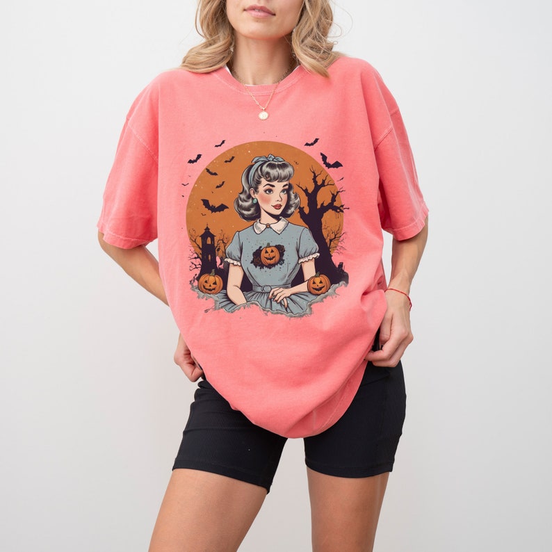 May include: A coral pink t-shirt with a vintage illustration of a woman in a blue dress with a pumpkin in her chest. The illustration is surrounded by bats and a full moon.