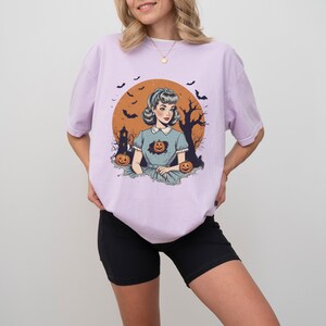May include: A lavender t-shirt with a vintage illustration of a woman in a blue dress with a pumpkin in her chest. The illustration is set against a background of a full moon, a spooky house, and bats.