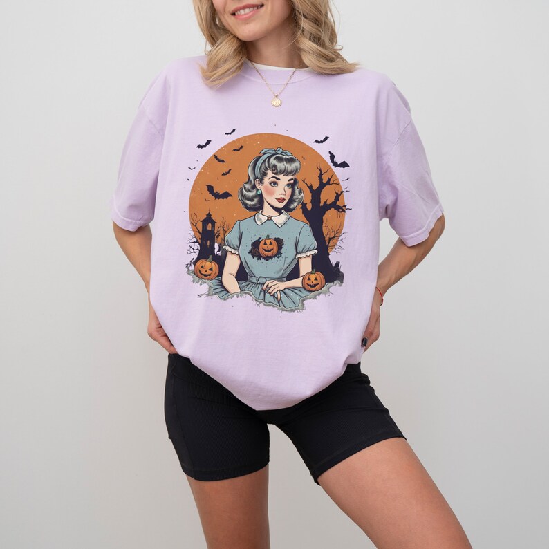 May include: A lavender t-shirt with a vintage illustration of a woman in a blue dress with a pumpkin in her chest. The illustration is set against a background of a full moon, a spooky house, and bats.