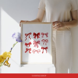 May include: A framed print with a white background and 12 red watercolor bows of various styles. The print is in a wooden frame and is sitting on a white surface.