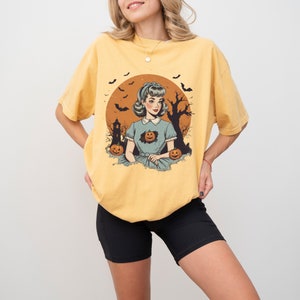 May include: A yellow t-shirt with a vintage illustration of a woman in a blue dress with a pumpkin in her chest. The illustration is set against a background of a full moon, bats, and a spooky tree.