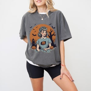 May include: A charcoal gray t-shirt with a vintage illustration of a woman in a blue dress holding a pumpkin. The illustration is set against a backdrop of a spooky forest and a full moon.