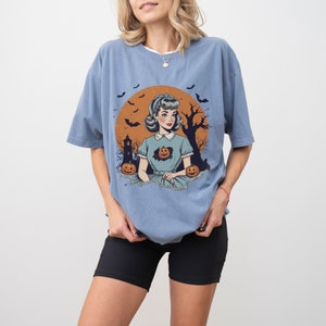 May include: A light blue t-shirt with a vintage illustration of a woman in a blue dress with pumpkins. The illustration is set against a background of a full moon, bats, and a spooky tree.