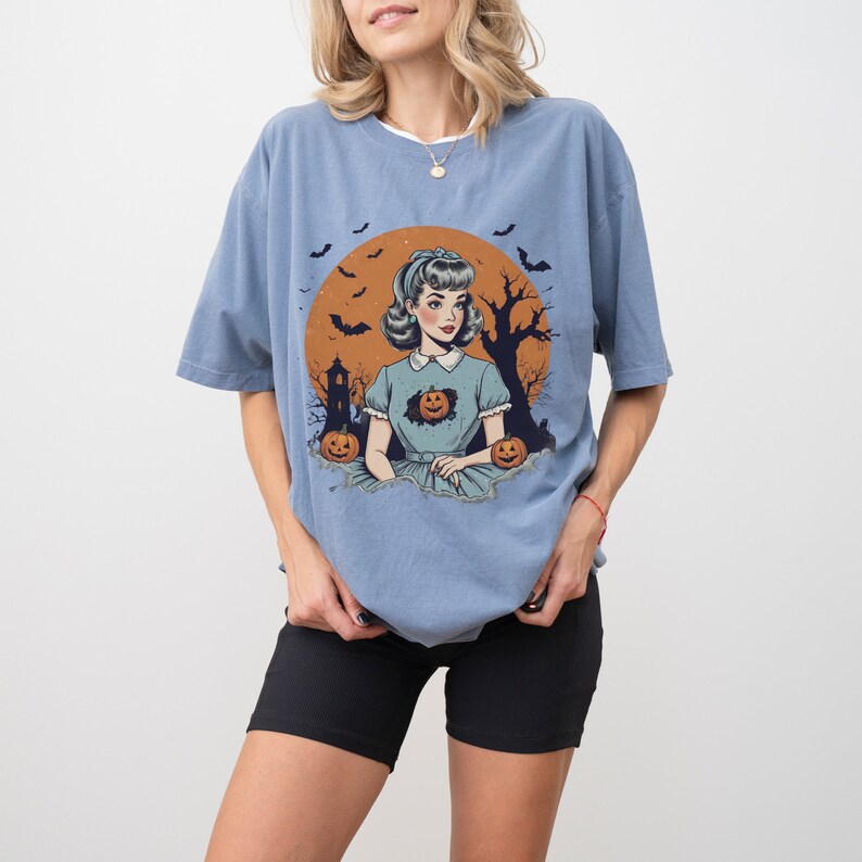 May include: A light blue t-shirt with a vintage illustration of a woman in a blue dress with pumpkins. The illustration is set against a background of a full moon, bats, and a spooky tree.