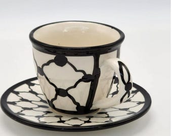 May include: A white ceramic teacup and saucer set with a black geometric pattern. The cup has a rounded shape with a handle, and the saucer is circular. The rim of the cup and saucer are outlined in black.