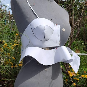 May include: A gray dress form with a white paper bra pattern pinned to it. The bra pattern has black lines and is held in place with red pins. The dress form is in a garden with yellow flowers.