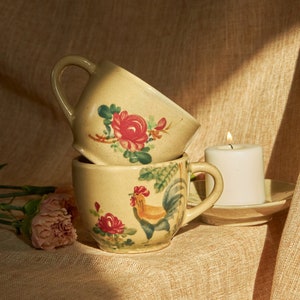 May include: Two ceramic mugs with floral and rooster designs stacked on top of each other. A white candle sits on a saucer next to the mugs.