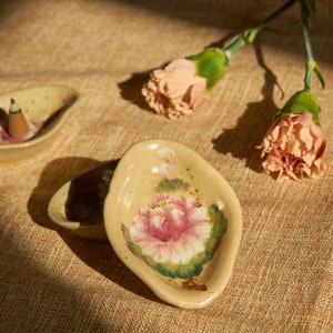 May include: Two small, ceramic incense holders with floral designs. One holder has a pink flower and green leaves, the other has a pink flower and green leaves with a small incense stick in the centre.