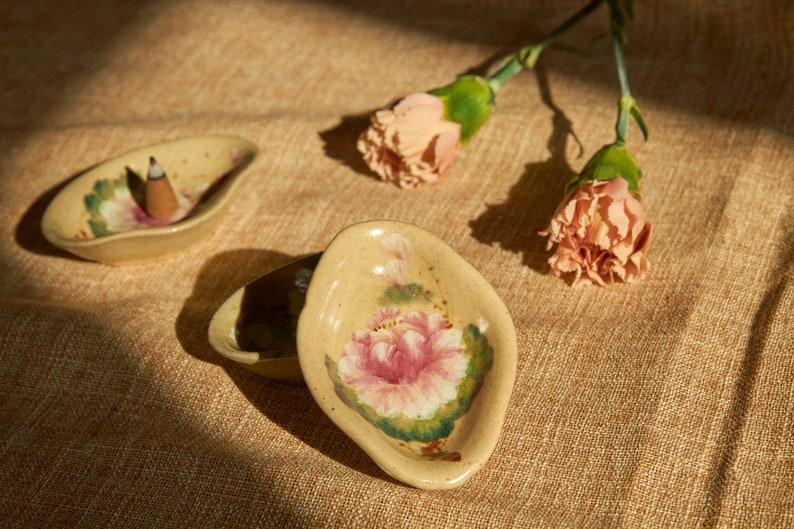 May include: Two small, ceramic incense holders with floral designs. One holder has a pink flower and green leaves, the other has a pink flower and green leaves with a small incense stick in the centre.