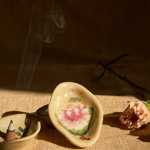 May include: Two ceramic incense holders with floral designs. One holder is a light brown colour with a pink flower and green leaves. The other holder is a darker brown colour with a green leaf design. A white incense cone is burning in the darker holder.
