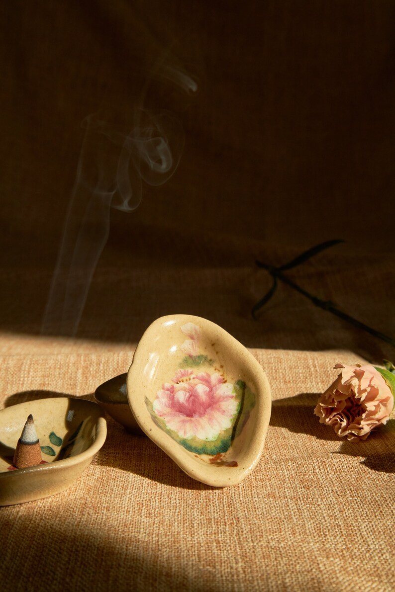 May include: Two ceramic incense holders with floral designs. One holder is a light brown colour with a pink flower and green leaves. The other holder is a darker brown colour with a green leaf design. A white incense cone is burning in the darker holder.