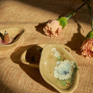 May include: Two small, ceramic incense holders with floral designs. The holder on the left has a cone-shaped incense stick inside. The holder on the right has a white flower design with green and brown accents.