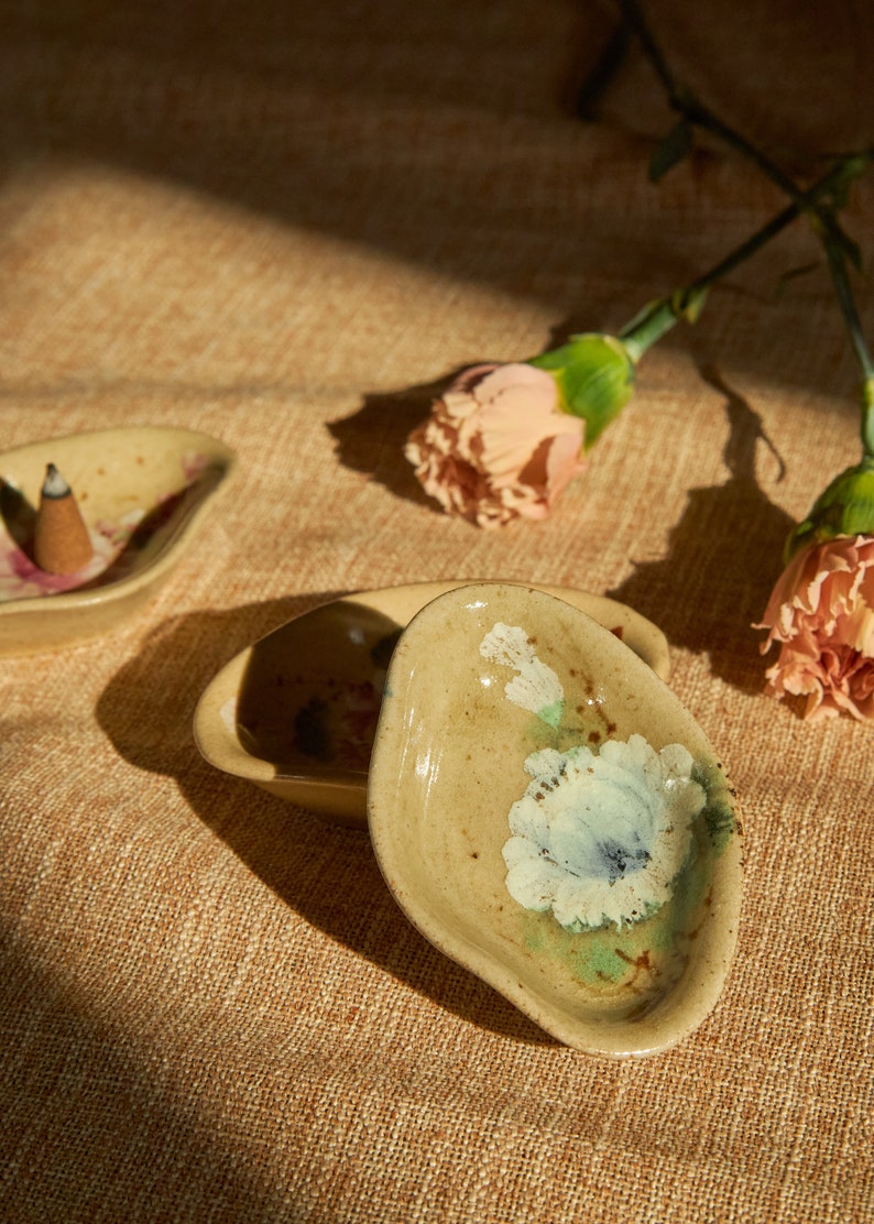 May include: Two small, ceramic incense holders with floral designs. The holder on the left has a cone-shaped incense stick inside. The holder on the right has a white flower design with green and brown accents.