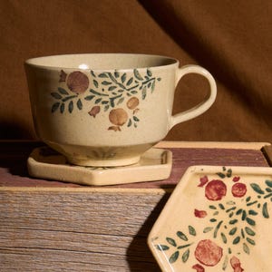 May include: A beige ceramic teacup and saucer with a floral design. The cup has a handle and the saucer is hexagonal. The design features green leaves and red pomegranate fruit.