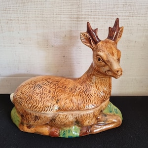 Könnte beinhalten: Eine Keramikdose mit Deckel in Form eines ruhenden Hirsches. Der Hirsch hat ein strukturiertes braunes Fell, dunkelbraunes Geweih und ruht auf einem Sockel, der mit grünen Grasdetails bemalt ist. Der Deckel trennt sich in der Körpermitte.