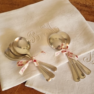 May include: A collection of silver spoons and forks tied with red and white ribbon, resting on a white embroidered linen cloth. The cloth features elegant embroidered initials. The spoons and forks are arranged in small bundles, perfect for a vintage-themed table setting.