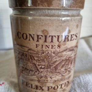 May include: A cylindrical brown ceramic jar with a glossy finish. The front features the text CONFITURES FINES and FELIX POTIN PARIS, along with a detailed brown graphic illustration of a factory landscape within two circular frames.