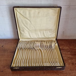 May include: A set of silver-toned spoons arranged in a rectangular dark brown storage case with a hinged lid. The interior of the case is lined with cream-coloured fabric, featuring individual slots to hold each spoon in a neat, parallel row.