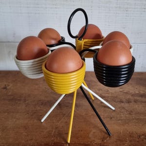 May include: A yellow, white and black wire egg holder with six egg cups. The holder has a circular base with three legs and a handle on top.