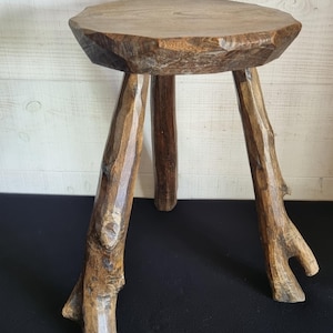 May include: A small, rustic wooden stool with a round seat and three branch-like legs. The stool is made of natural wood with visible grain and knots, and has a warm brown colour. The legs are angled outwards for stability.