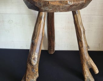 Puede incluir: Un pequeño taburete de madera rústica con un asiento redondo y tres patas en forma de rama. El taburete está hecho de madera natural con vetas y nudos visibles, y tiene un color marrón cálido. Las patas están anguladas hacia afuera para mayor estabilidad.