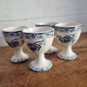 May include: A set of four cream-colored ceramic egg cups featuring a dark blue floral pattern around the rim and base. Each cup has a pedestal foot and a rounded bowl shape, decorated with detailed blue botanical illustrations on the exterior surface.