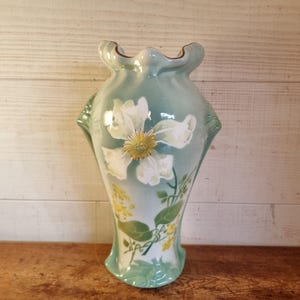 Könnte beinhalten: Eine Vintage-Keramikvase mit hellblauem und weißem Blumendesign. Die Vase hat eine große weiße Blume mit gelbem Zentrum, grüne Blätter und gelbe Akzente. Die Vase hat einen gerüschten Rand und steht auf einer Holzoberfläche.