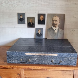 Puede incluir: Una caja de madera vintage con una tapa negra y asas de latón. La tapa tiene una etiqueta que dice "Couples regard et factures. Rituel des Chapels". Hay cinco pequeños retratos enmarcados en la pared detrás de la caja.