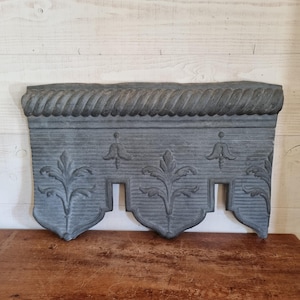 Op de afbeelding: Een grijs, decoratief architecturaal element met een touwachtige rand en gestileerde bloemmotieven. Het stuk heeft drie hangerachtige secties met een centrale uitsparing. Het item is gemaakt van een solide materiaal, mogelijk steen of beton.