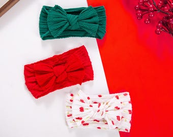 Puede incluir: Tres diademas con lazos. Una es verde, otra roja y la tercera blanca con un estampado de cerezas rojas. Las diademas son de tela texturizada y están sobre un fondo blanco y rojo.