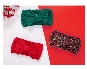 Puede incluir: Tres diademas para bebés en rojo, verde y burdeos estampado. Cada diadema presenta un lazo grande. Las diademas se muestran sobre un fondo rojo y blanco, con bayas rojas en la esquina superior izquierda.