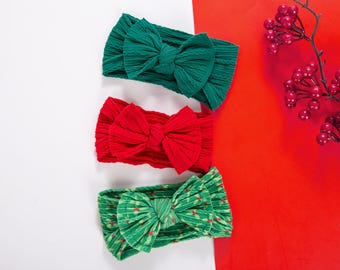 Puede incluir: Tres diademas para bebés en colores festivos. La diadema superior es verde, la del medio es roja y la inferior es verde con un estampado. Cada diadema tiene un lazo grande. El fondo es rojo y blanco.