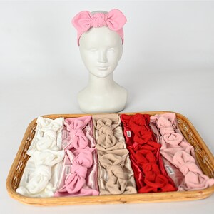 May include: A wicker basket filled with a variety of knotted headbands in different colors, including white, pink, beige, and red. The headbands are arranged in rows, with each row featuring a different color.