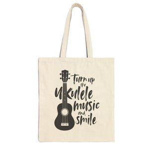 Puede incluir: Bolso tote beige con una ilustración de ukelele negro y el texto "Turn up the Ukulele music and smile." El bolso tiene asas largas, adecuado para llevar objetos.
