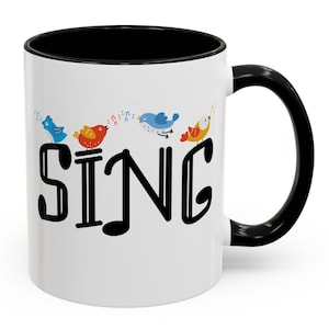 May include: White ceramic mug with a black interior, handle, and rim. The mug features the word "SING" in large black letters, with colorful cartoon birds above the letters. The birds are singing musical notes.