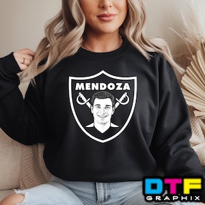 Könnte beinhalten: Schwarzes Sweatshirt mit einem weißen Schilddesign. Das Schild zeigt den Namen "MENDOZA" über einem Porträt und gekreuzten Schwertern. Das Sweatshirt hat einen Rundhalsausschnitt und lange Ärmel. In der unteren rechten Ecke steht der Text "DTF GRAPHIX" in Blau, Gelb und Rosa.
