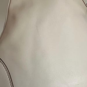 May include: A close-up of a white leather handbag with a smooth texture. The bag has a curved shape and a stitched edge.