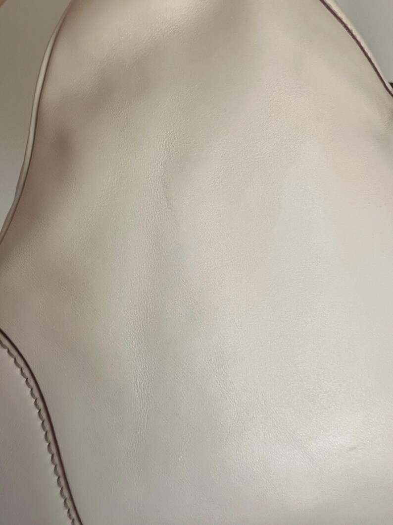 May include: A close-up of a white leather handbag with a smooth texture. The bag has a curved shape and a stitched edge.